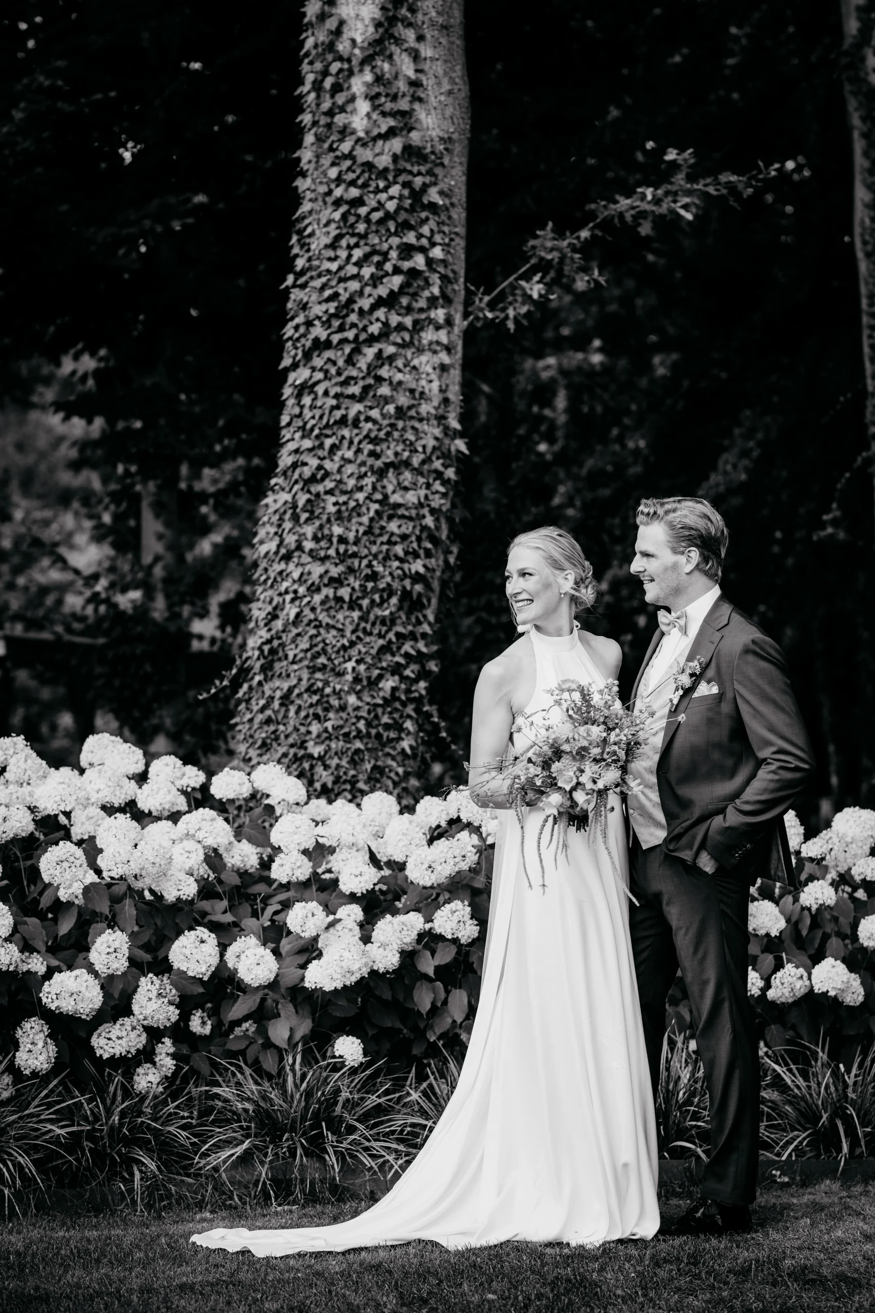 Ein Hochzeitspaar auf einem Gartenweg unter Bäumen. Schwarz-Weiß-Fotografie.