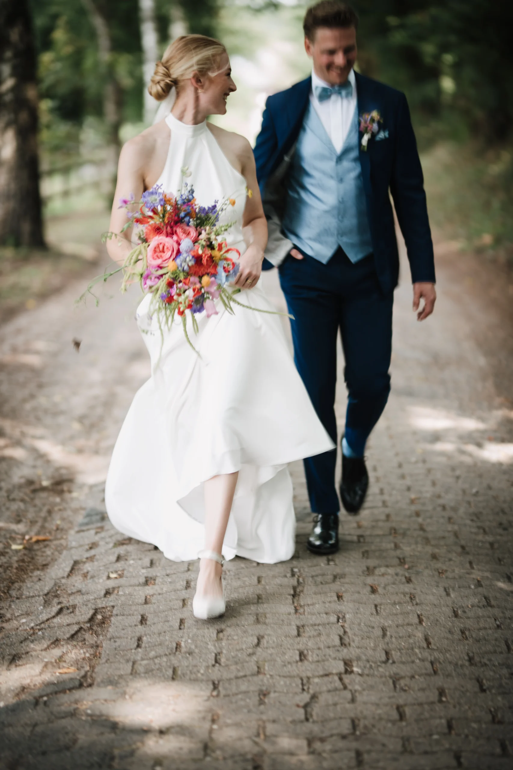 Die Braut hält ihren Blumenstrauß und ihr Kleid, während das Hochzeitspaar auf einem Weg geht.