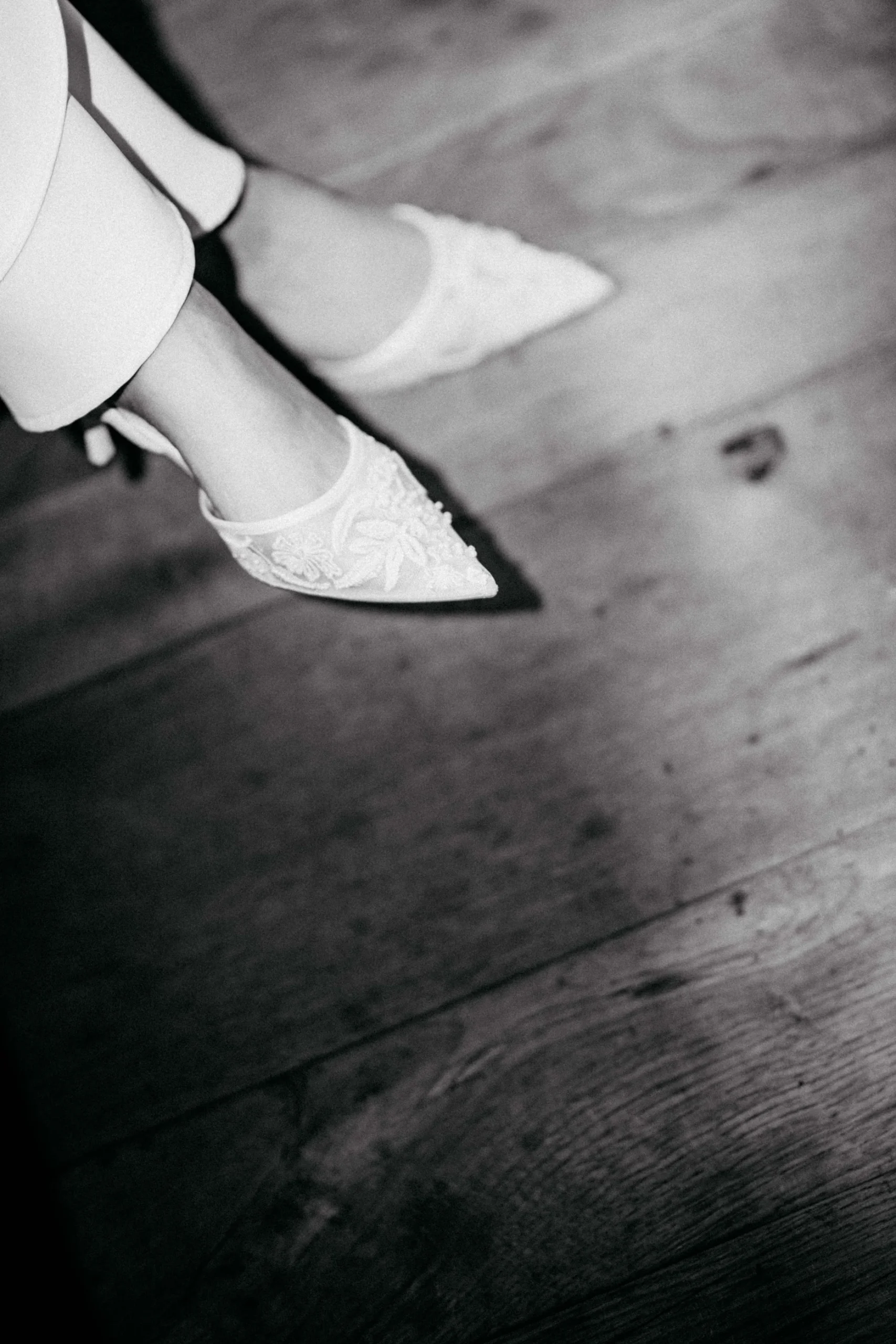 Weiße Brautschuhe unter dem Saum eines Kleides auf einem Holzboden. Schwarz-Weiß-Fotografie.
