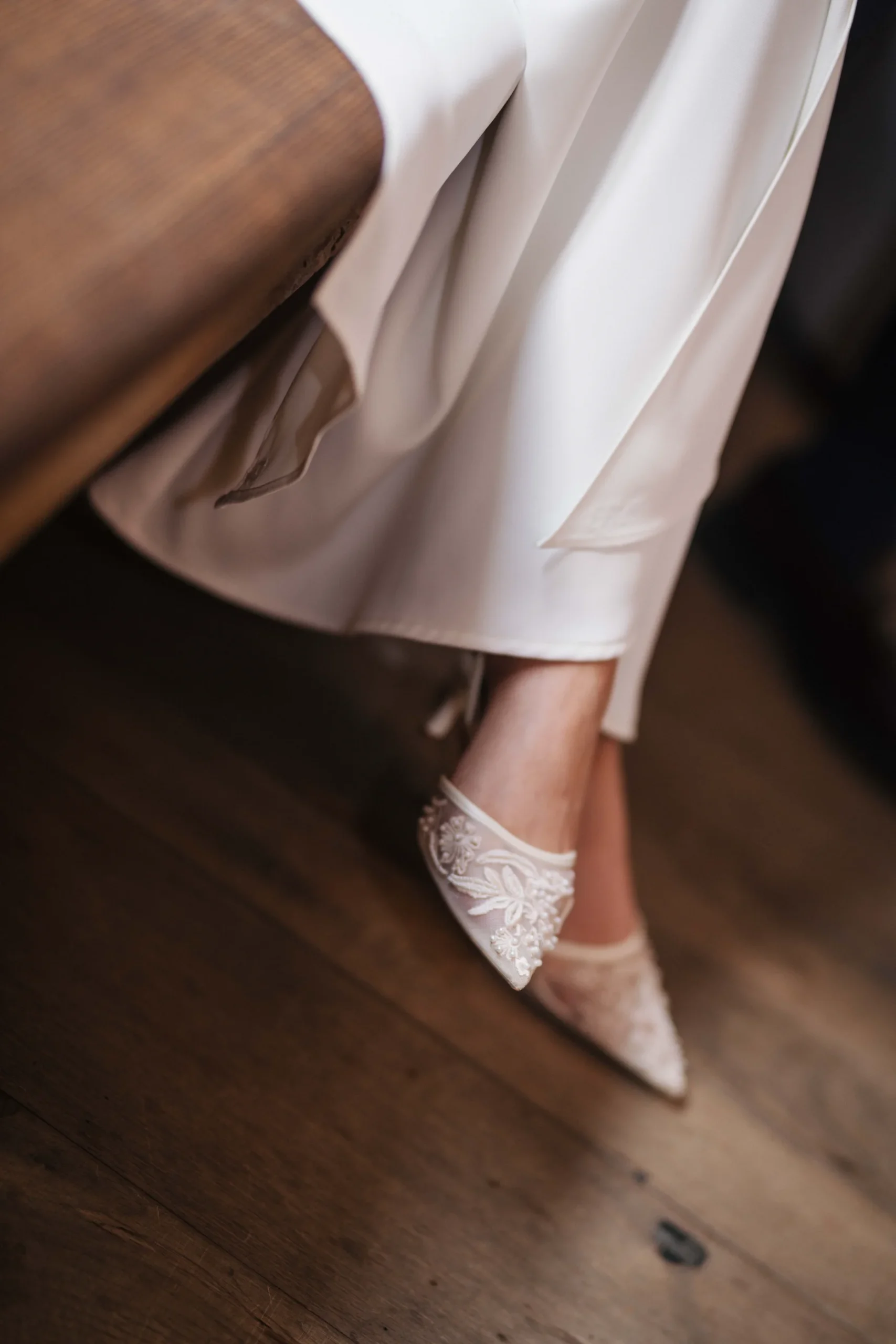 Ein heller Brautschuh mit Schmuckdetails unter einem langen Kleid neben einer Holzbank.