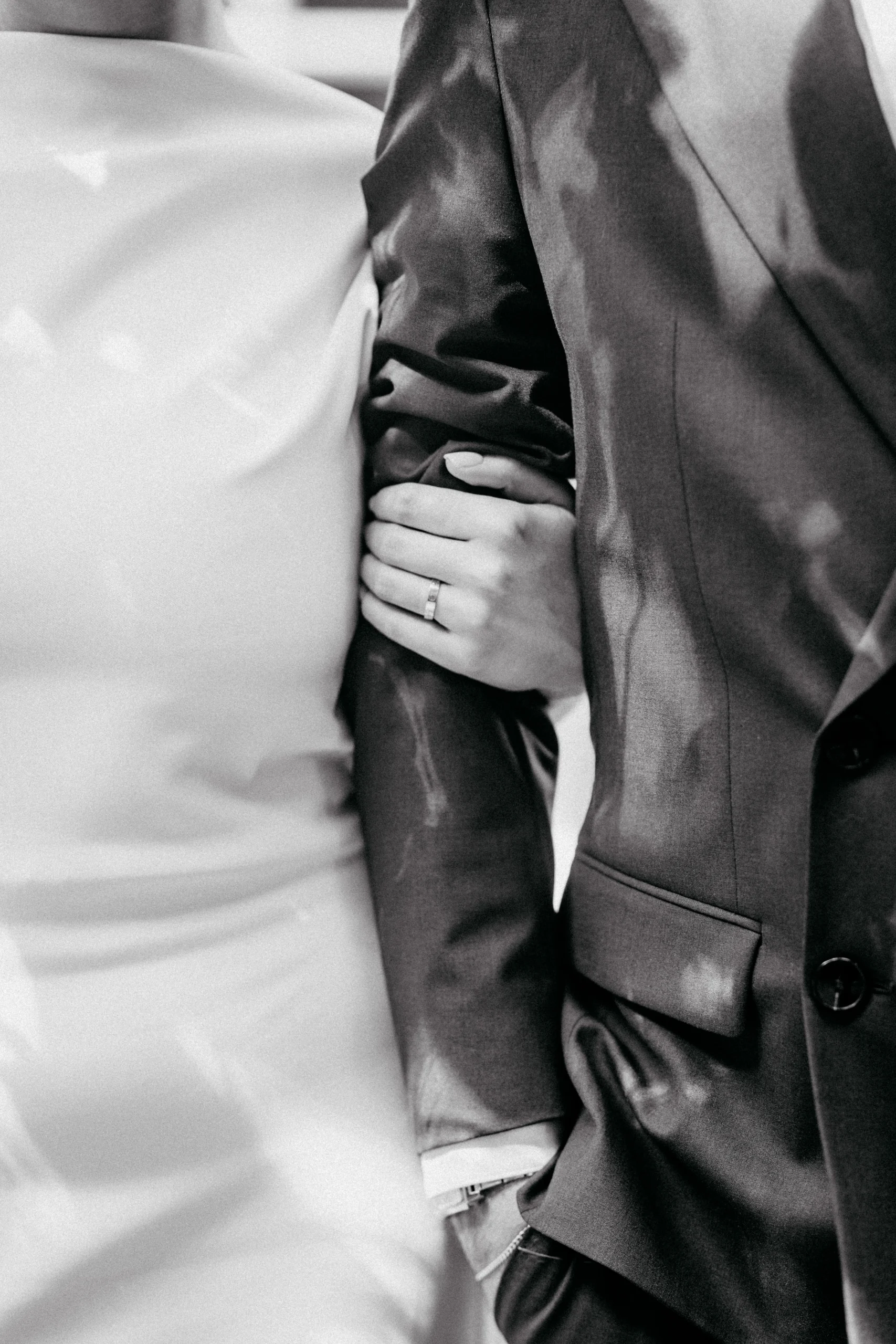 Nahaufnahme einer Hand mit Ehering am Arm eines dunklen Anzugs. Schwarz-Weiß-Fotografie.