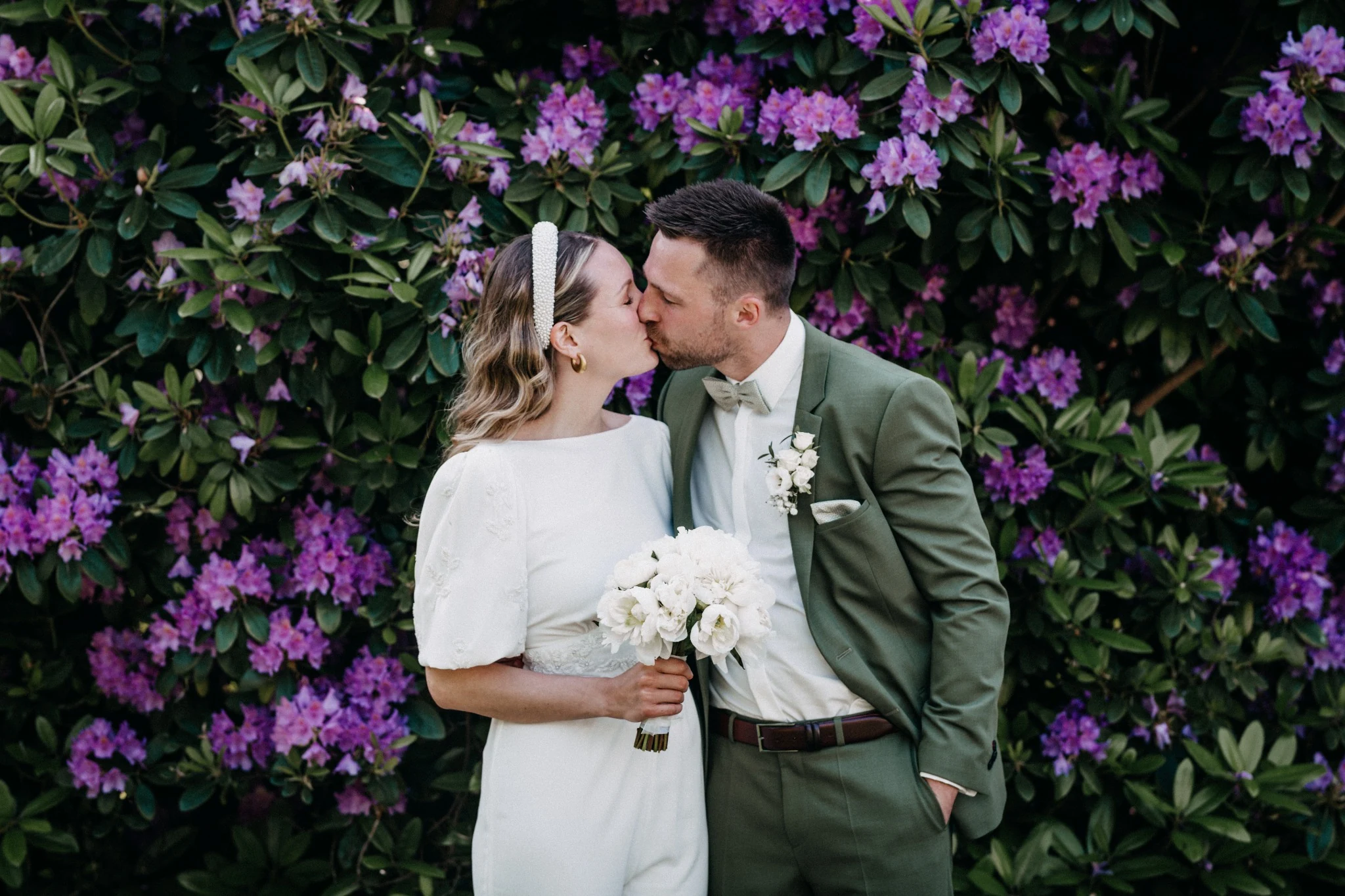Birthe und Niklas küssen sich vor violett blühenden Rhododendren. Birthe trägt einen weißen Jumpsuit, Niklas einen grünen Anzug.