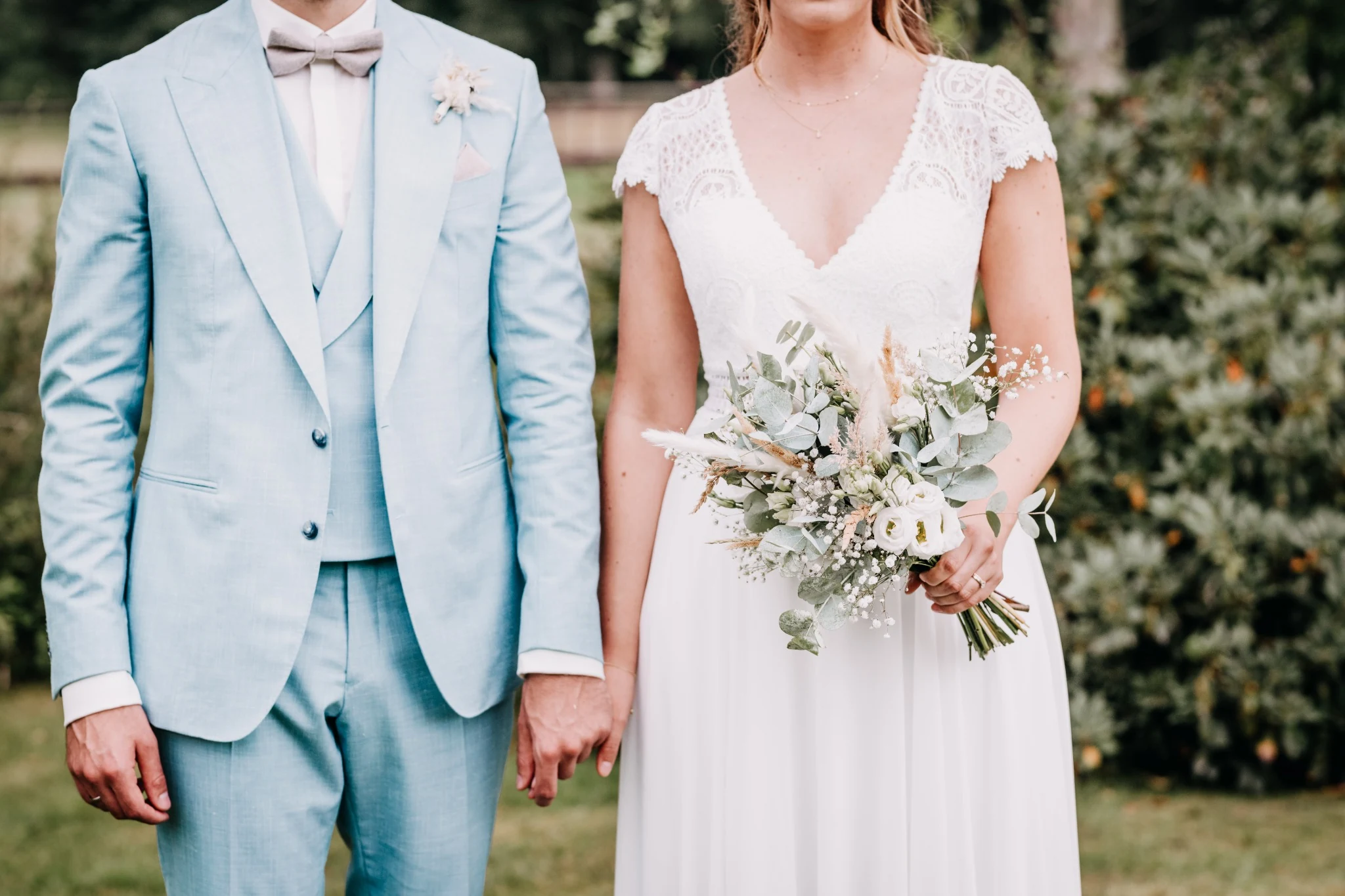 Desi und David halten sich an den Händen. Der Bildausschnitt zeigt den hellblauen Anzug, das weiße Spitzenkleid und den Brautstrauß.