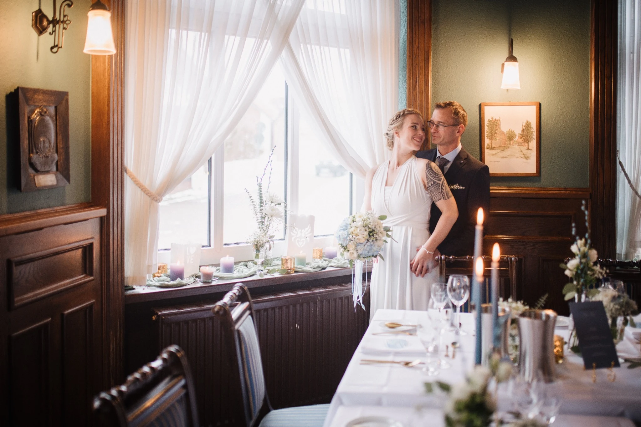 Ronja und Markus stehen am hellen Fenster im Hotel Altes Land, umgeben von Kerzen und gedeckten Tischen.