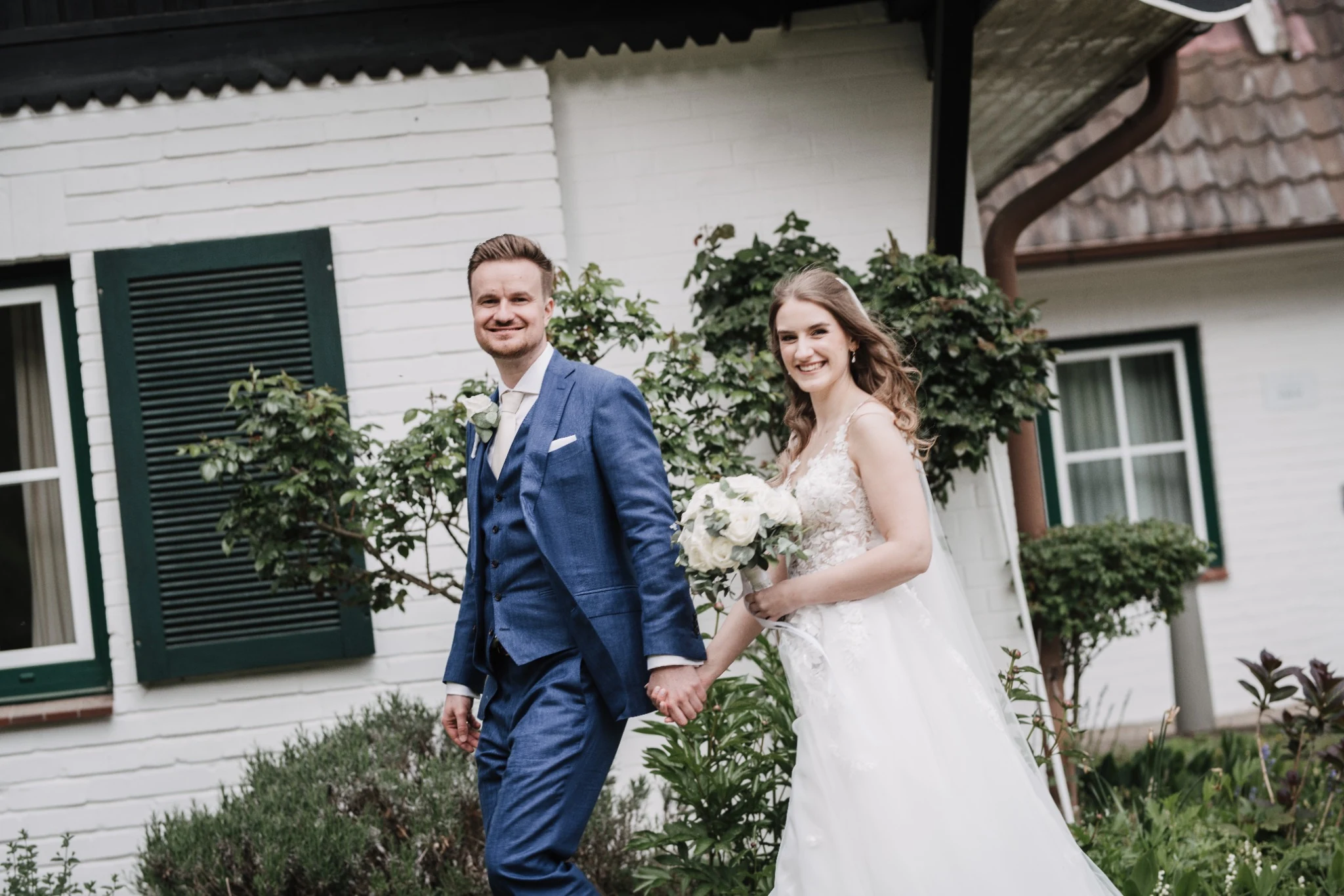 Shannon und Philipp gehen lachend am weißen Haus mit grünen Fensterläden vorbei.