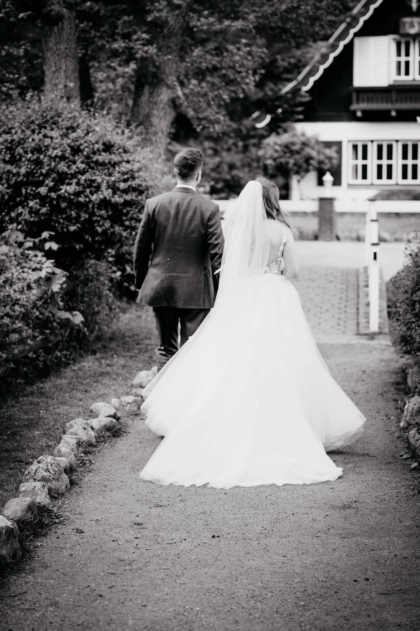 Shannon und Philipp gehen gemeinsam einen Weg entlang auf ein Fachwerkhaus zu. Der lange Schleier und das Brautkleid sind von hinten zu sehen. Schwarz-Weiß-Fotografie.